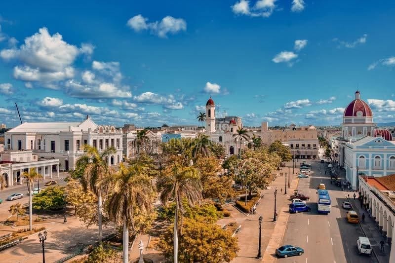 A vibrant urban scene unfolds with a wide avenue lined by palm trees. Historical buildings with varied architectural styles surround a central plaza, while a few vehicles are parked along the street. Bright blue skies and scattered clouds create a lively backdrop.