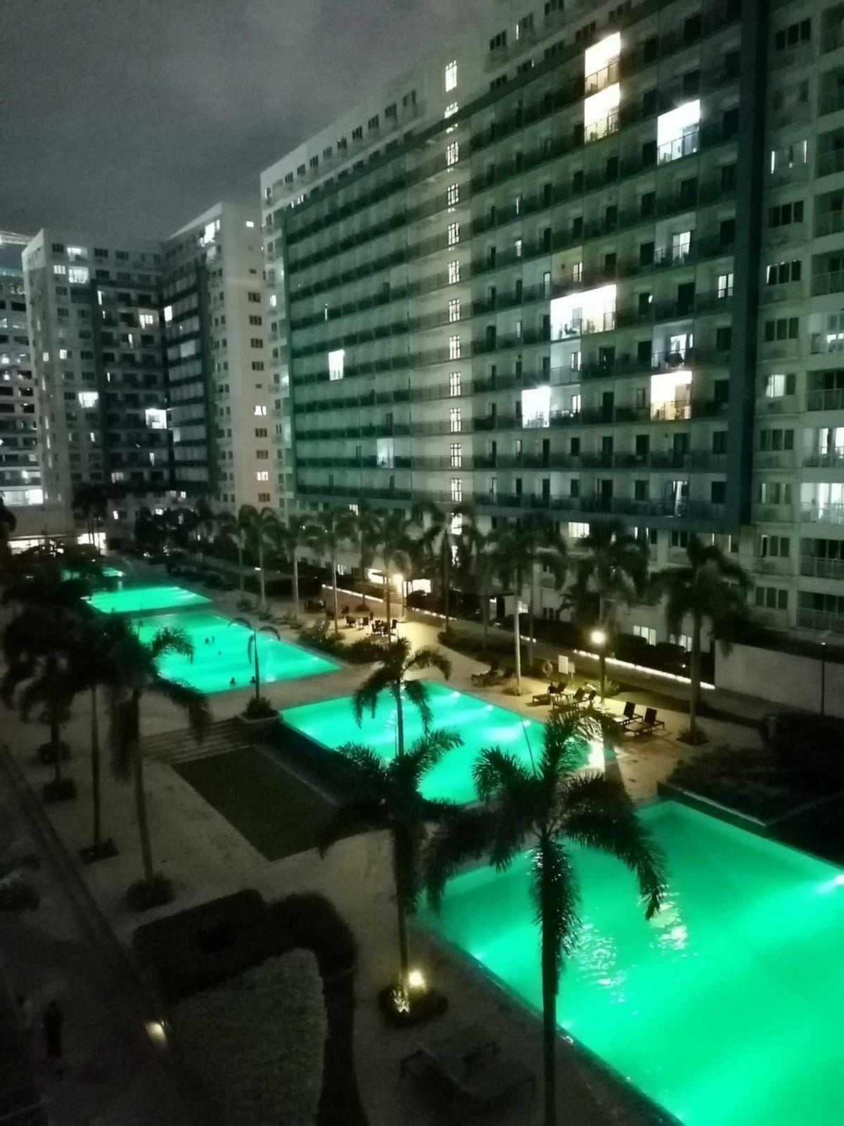 A nighttime view of the pool area showcases multiple illuminated swimming pools, each glowing in turquoise tones. Surrounding palm trees provide a tropical ambiance, while the high-rise buildings display warm lights in the windows. The scene conveys a relaxed outdoor space for guests.