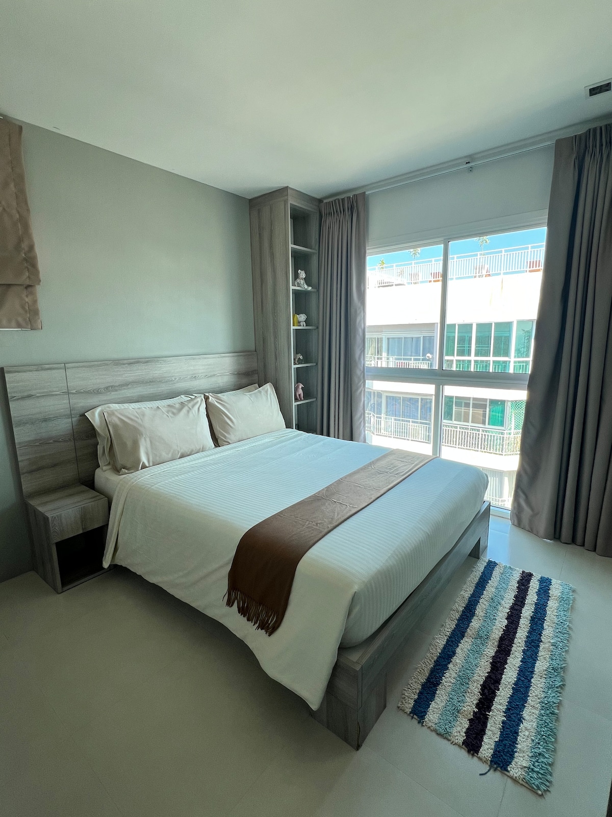 A cozy sleeping area features a spacious bed with light-coloured linens and decorative pillows. A smooth wooden headboard complements the neutral walls. Natural light floods the room through large windows draped with grey curtains, creating a peaceful ambiance.