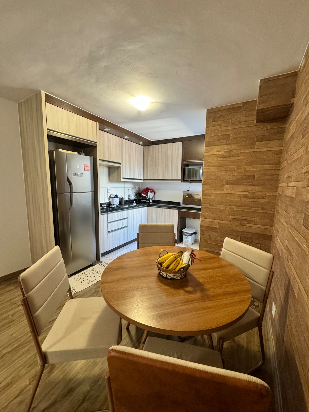 A compact kitchen space features wooden cabinetry and modern appliances, including a stainless-steel refrigerator and microwave. A round wooden table is surrounded by four chairs. A basket of bananas is placed at the center of the table, enhancing the kitchen's functionality.