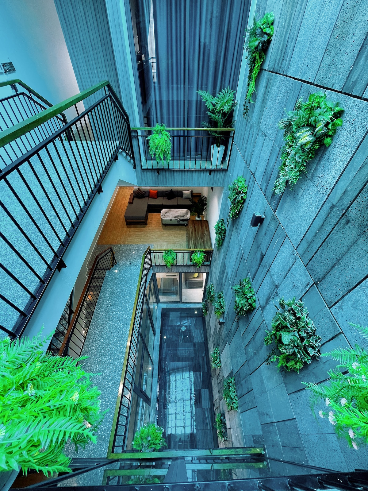 An aerial view of an interior courtyard is presented, featuring a blend of greenery and modern design. Vertical planters adorn the stone walls, while a glimpse of a spacious living area is visible below. Metal railings encircle the open space, enhancing the inviting atmosphere.