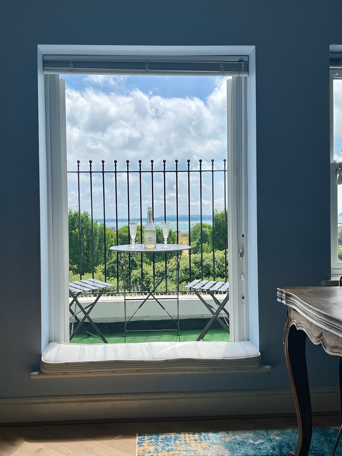 A large window frames a delightful outdoor balcony scene. A small bistro table with two chairs is set with bottles, offering a view of the sea and greenery beyond. Natural light filters in, highlighting the inviting interior space.