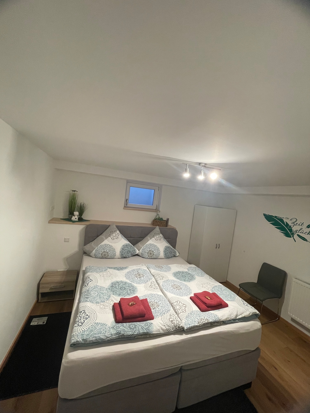 A well-lit bedroom features a spacious bed with patterned bedding. Two red towels are neatly arranged on top. A small plant sits on a shelf above the headboard. A chair and a closet are also visible, completing the functional layout of the space.