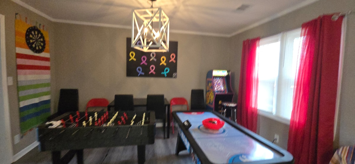 A recreational room features a foosball table and an air hockey table, both positioned alongside six black and red chairs. A colorful dartboard and a retro arcade game are visible, while large red curtains frame the windows, allowing natural light to brighten the space.