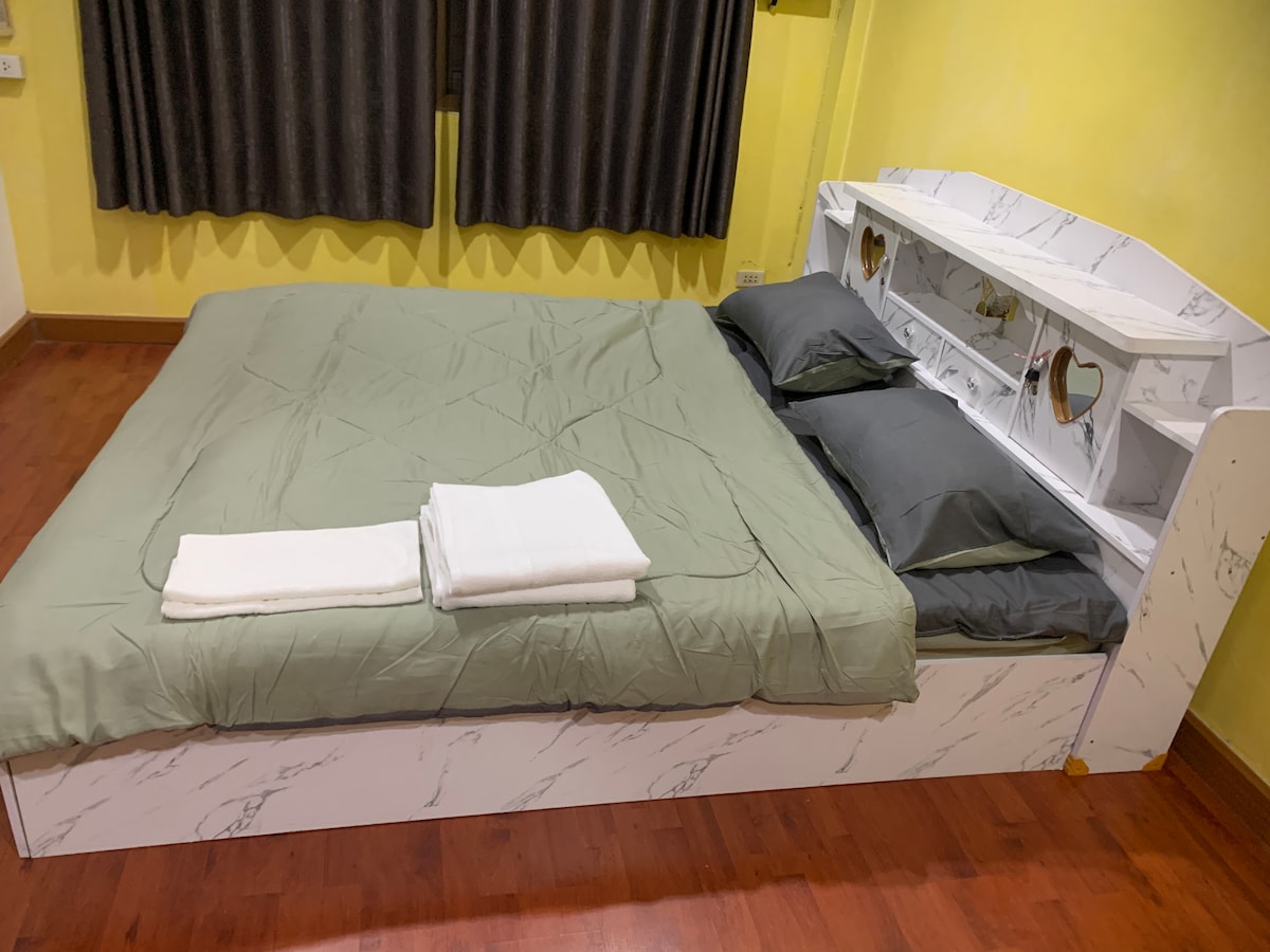 A spacious bed is positioned against a yellow wall, featuring a marbled headboard. Soft gray and green bedding is neatly arranged, with white towels placed at the foot of the bed. Dark curtains frame the window, complementing the warm interior.