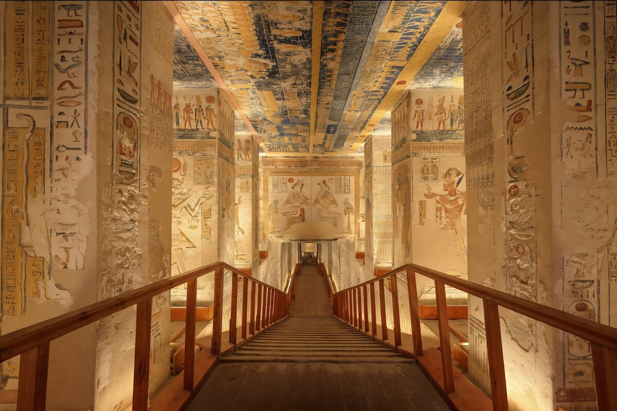 A grand staircase leads down into a richly decorated chamber featuring vibrant hieroglyphics and intricate wall paintings. The ceiling is adorned with detailed artwork in shades of blue, creating a striking contrast with the warm illumination provided by carefully positioned lights.