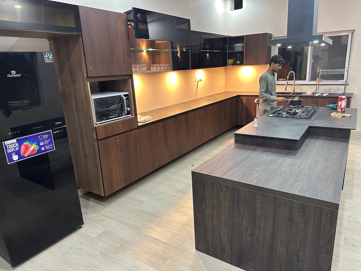 A modern kitchen is shown, featuring sleek wooden cabinetry and a central island. Stainless steel appliances, including a refrigerator and a gas stove, are visible. Soft lighting highlights the clean lines and spacious countertop, creating an inviting area for meal preparation and gatherings.