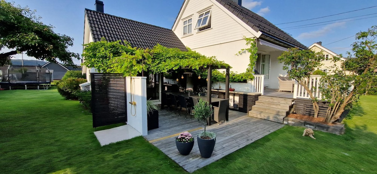 An outdoor dining area is featured under a pergola draped with greenery. A wooden deck with a large table and several chairs is present, complemented by potted plants. The house's exterior displays a combination of white siding and dark roofing, surrounded by a well-maintained lawn.