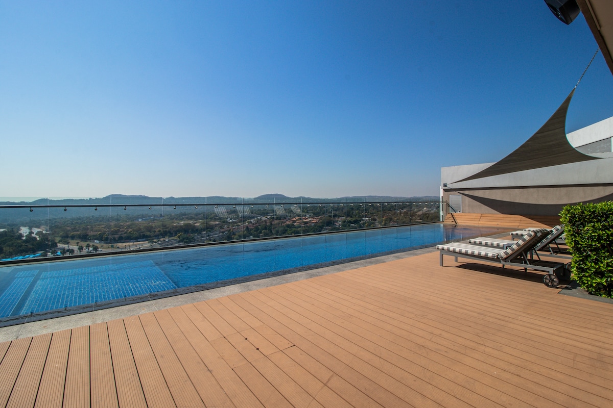 A serene rooftop area features an infinity pool with panoramic views of the surrounding landscape. Sun loungers are positioned beside the pool, set on a wooden deck, providing a relaxing space to enjoy the outdoors under clear blue skies.