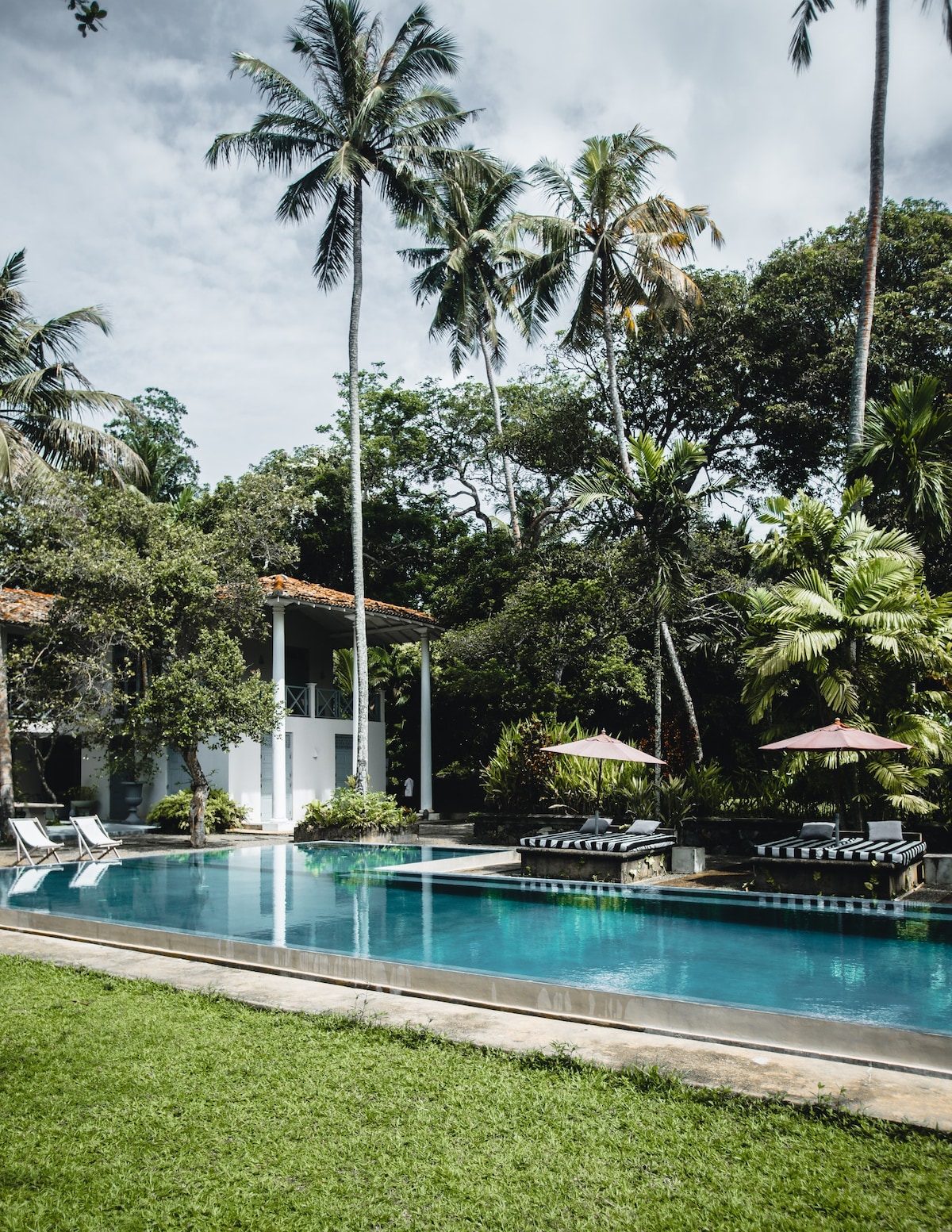 A serene swimming pool is surrounded by lush greenery, with tall palm trees providing shade. Sun loungers line one side of the pool, while patio umbrellas offer additional cover. The two-story villa is partially visible in the background, complementing the tranquil outdoor setting.