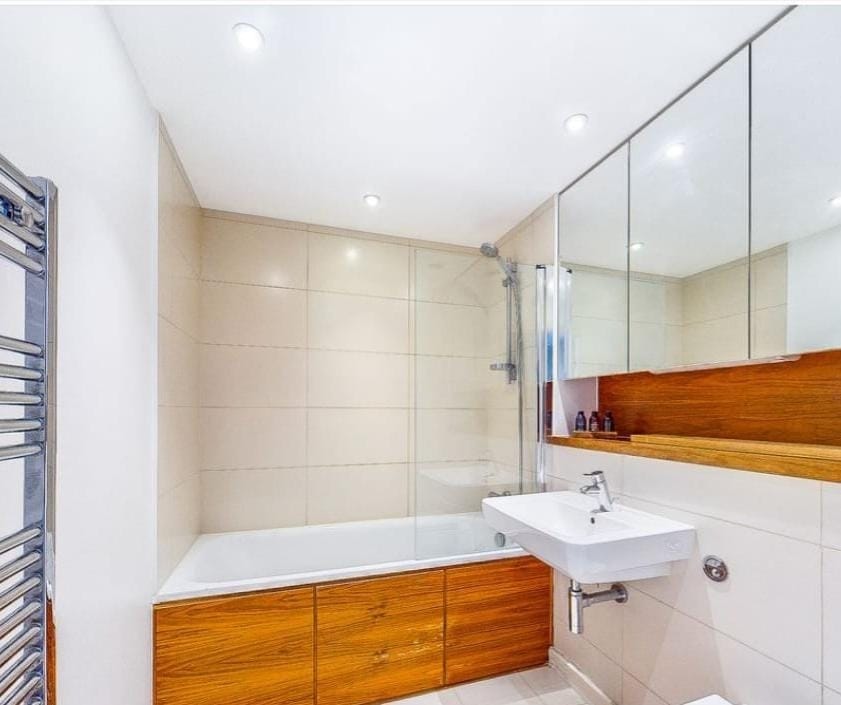A modern bathroom features a sleek, glass-enclosed shower and a bathtub with wood cabinetry below. A minimalist sink is positioned against the wall under a large mirror, and warm lighting highlights the neutral tile and wood finishes throughout the space.