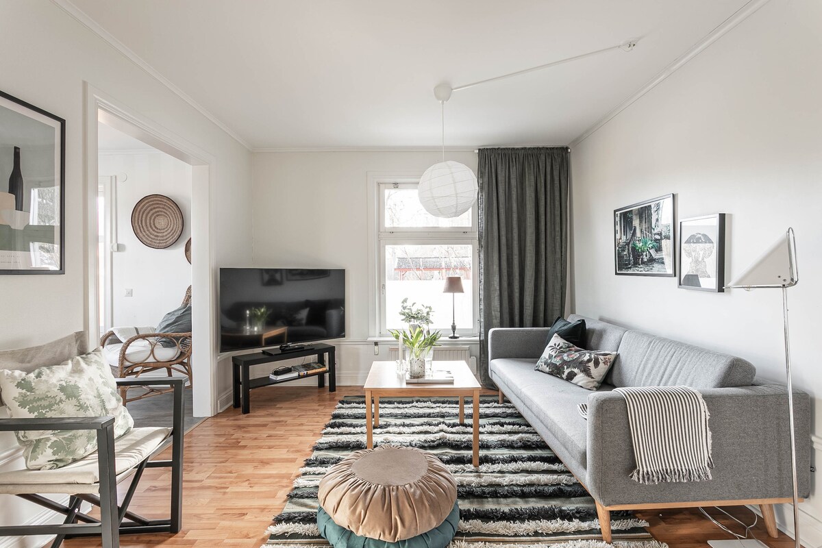 A comfortable living area is displayed, featuring a grey sofa paired with decorative pillows. A wooden coffee table rests on a striped rug, complemented by a round pouf. Natural light enters through large windows, casting a soft glow on the space. A television is positioned nearby.