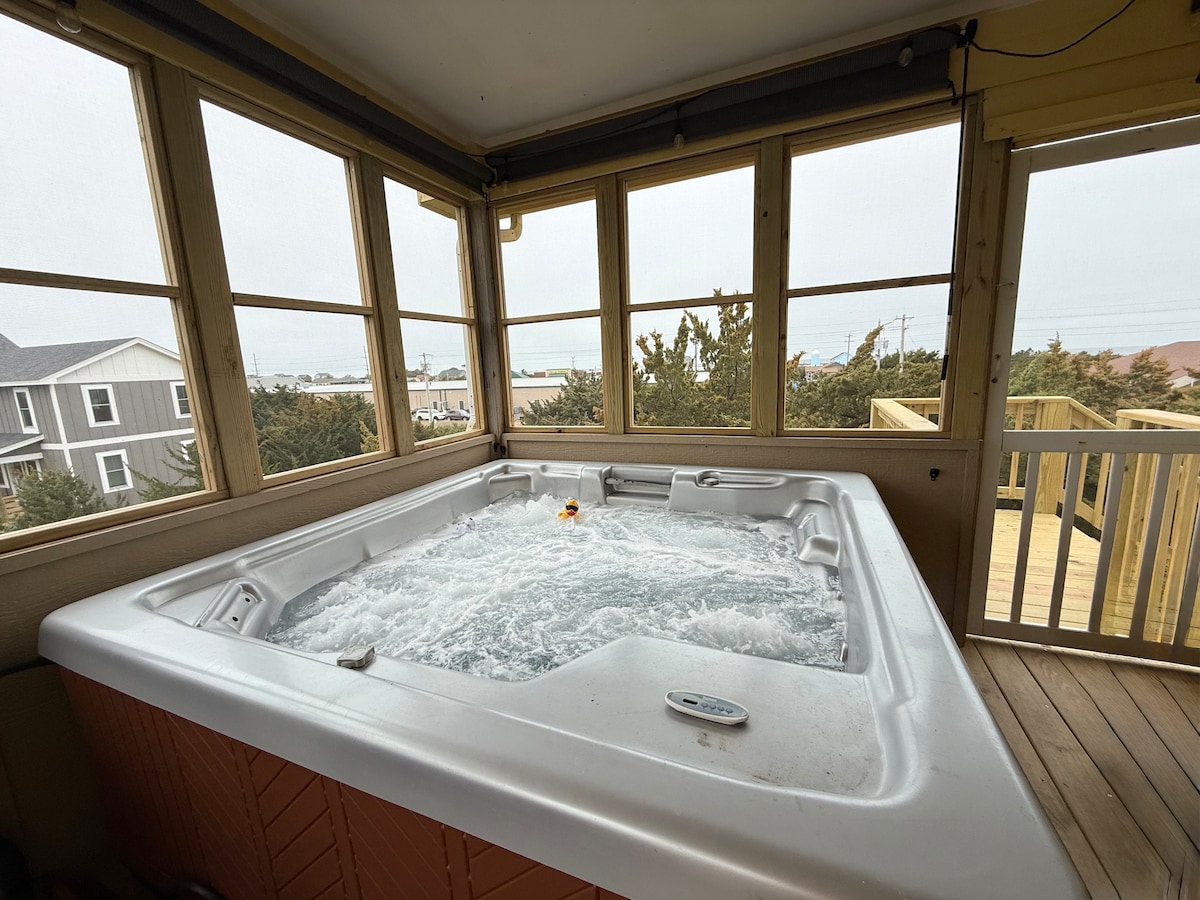 An indoor hot tub is situated in a sunlit room, featuring large windows offering views of the surrounding landscape. The bubbling water of the hot tub creates a calming ambiance. Wooden flooring extends throughout the area, complementing the natural light from the outside.