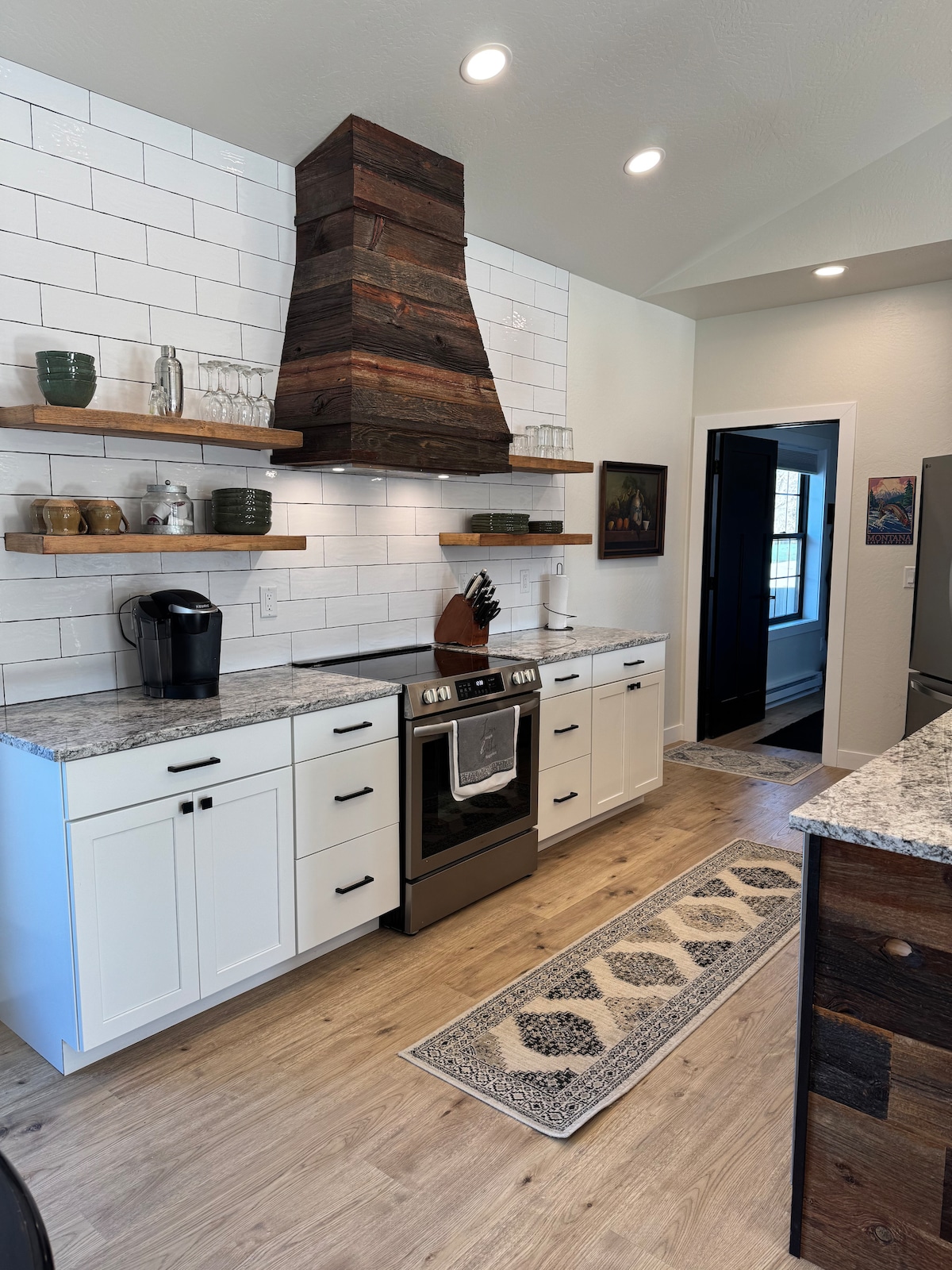 The modern kitchen features light cabinetry and dark wooden shelves against a tiled backsplash. A stainless steel range and vent are centrally positioned, while a coffee maker is placed nearby. A decorative runner extends along the wood floor, leading to a doorway.
