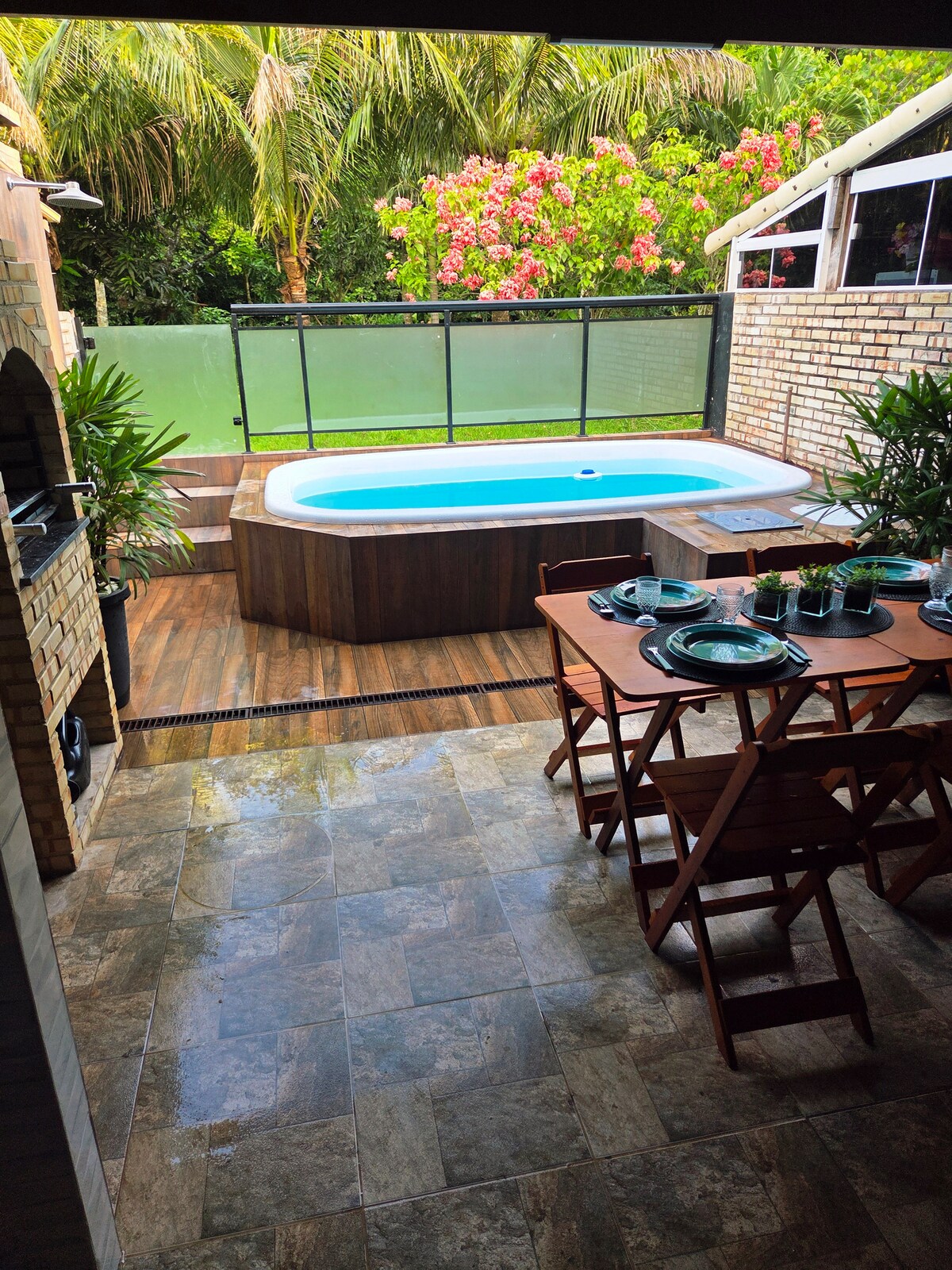 An outdoor area features a private swimming pool surrounded by greenery and flowering plants. A dining table with plates and utensils is set nearby. The space includes tiled flooring and a wooden deck, providing a cozy setting for relaxation and meals.