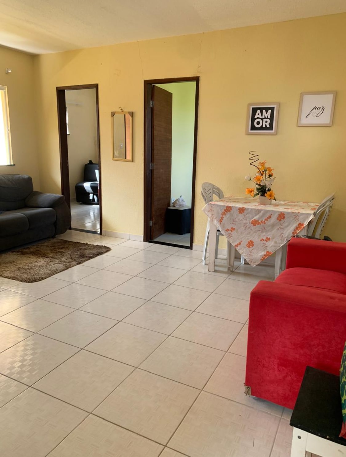 A cozy living area is presented with a mix of furniture, including a dark sofa and a bright red chair. A small table adorned with a floral tablecloth and a vase of flowers is positioned nearby, while doorways lead to connecting rooms.