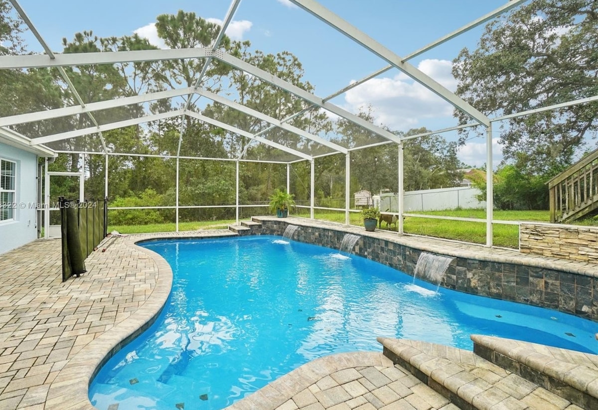 A custom pool with jets is set within a spacious, fully enclosed lanai. The clear blue water reflects natural light, and stone accents frame the pool area. Waterfalls tumble gently into the pool, surrounded by lush greenery and a well-maintained backyard.