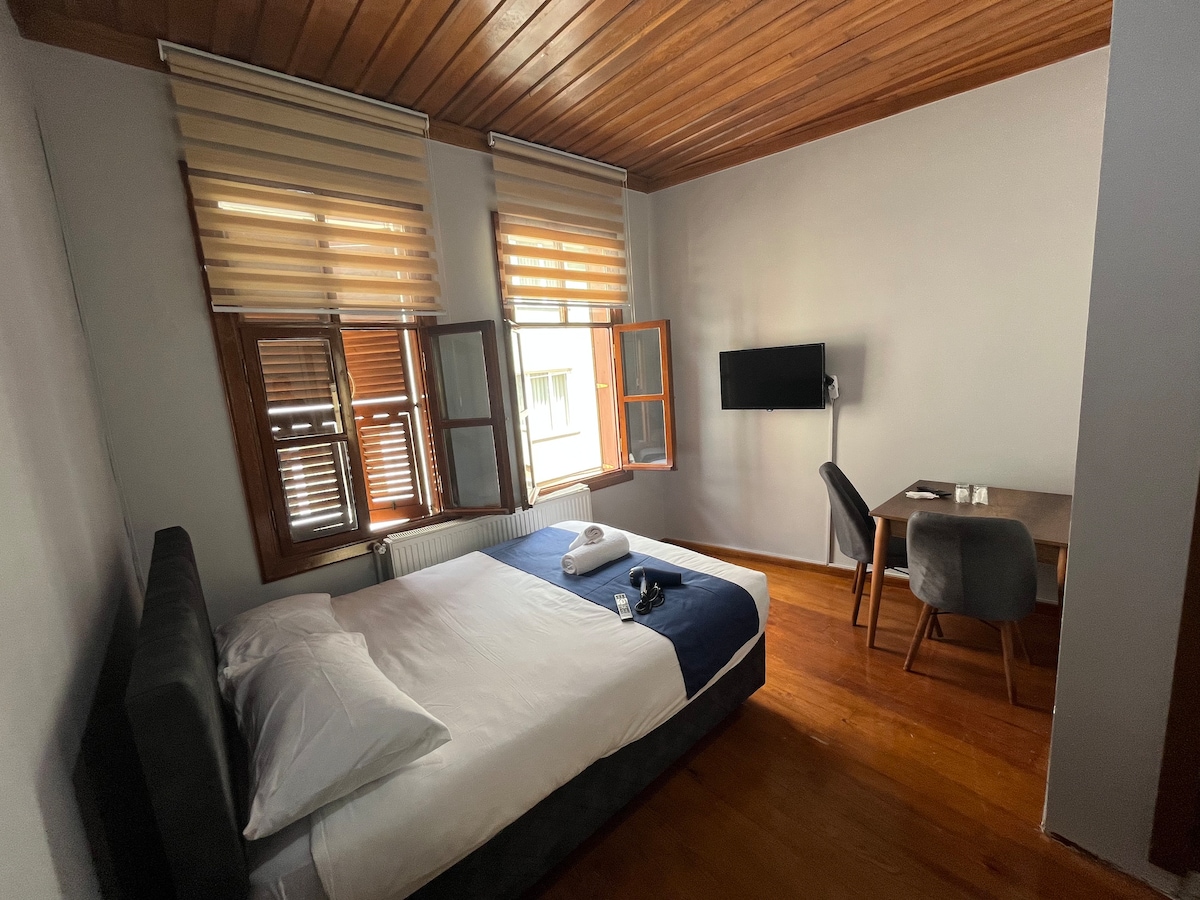 A bright room features a spacious bed with fresh linens and towels. Two windows are equipped with wooden shutters, allowing natural light to fill the space. A small table and chair are positioned nearby, and a wall-mounted TV adds convenience.