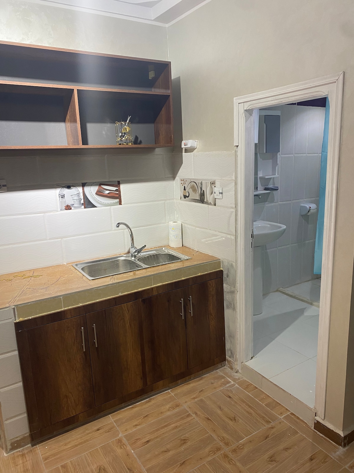 A compact kitchen area is presented, featuring a utilitarian sink and cabinetry below. Open shelving above holds a few items. A doorway on the right leads to a bathroom, which includes light-colored tiles and visible fixtures.
