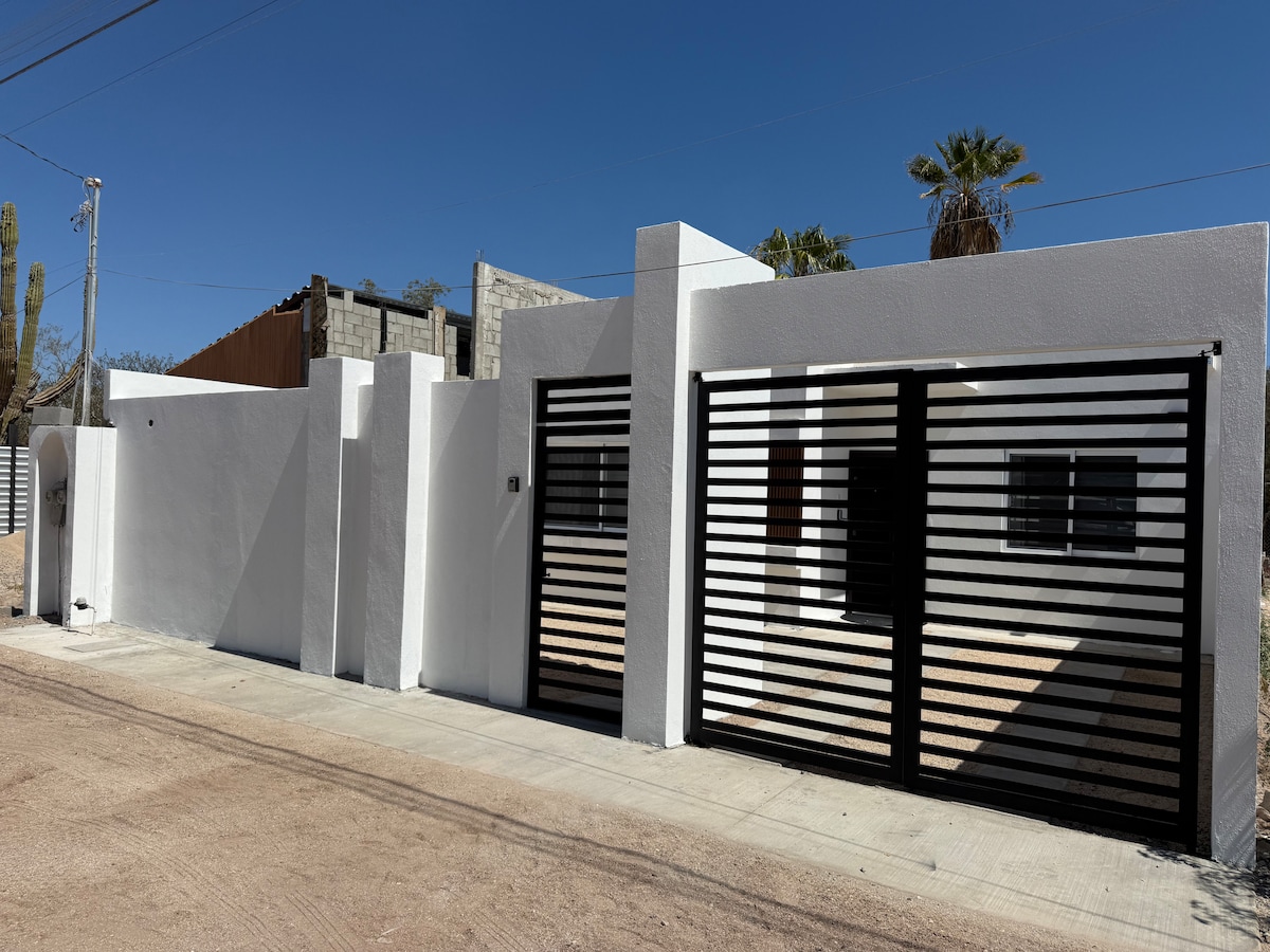 A modern architectural facade is presented, featuring a combination of white walls and black horizontal metal accents. The entrance is framed by a gate and a low wall. The surrounding landscape includes sparse desert vegetation, indicative of a sunny, dry climate.