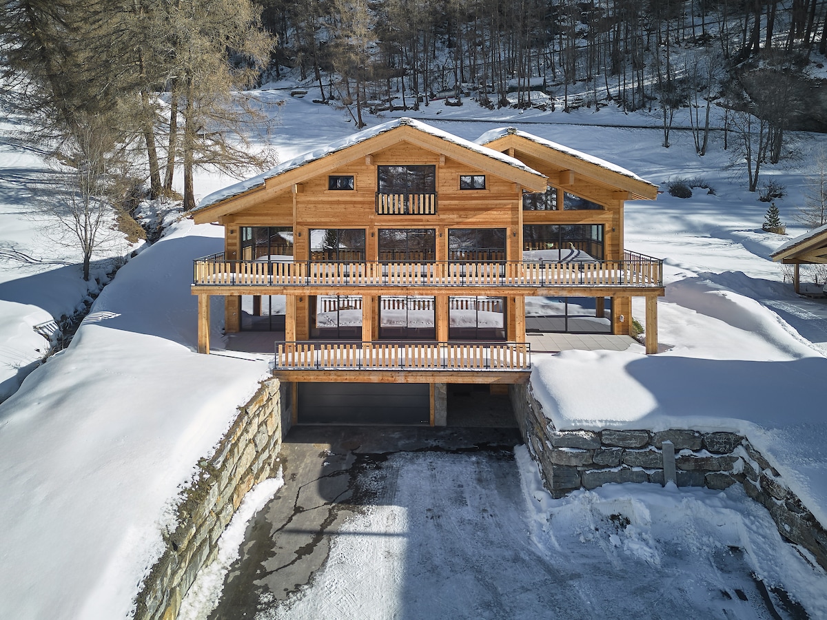 A spacious chalet is nestled in a winter landscape, featuring a warm wooden exterior and multiple balconies. Snow-covered ground surrounds the structure, and large windows provide a view of the serene environment. A parking area is visible beneath the elevated building.
