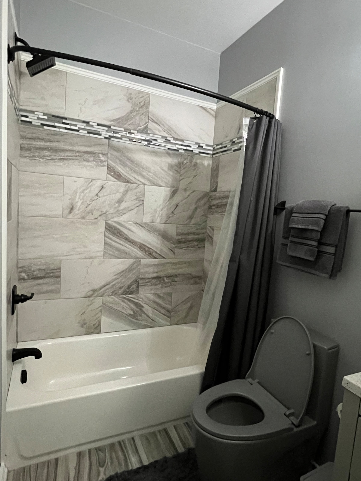 A modern bathroom features a bathtub with a shower curtain, complemented by sleek gray-toned walls adorned with textured marble tiles. A toilet sits nearby, and gray towels are neatly arranged on a wall-mounted rack.