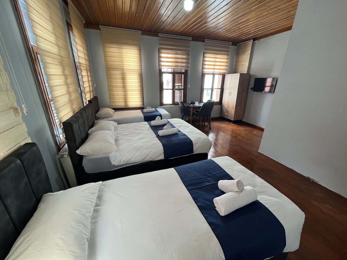 A spacious guestroom features three beds with white linens and dark blue accents. Light filters through multiple windows adorned with wooden blinds, enhancing the warm wooden ceiling. A small dining table and a wall-mounted television are also visible in the inviting space.