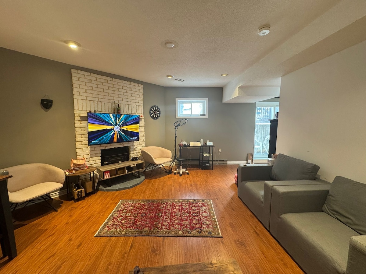 A light-filled living area features a flat-screen television mounted above a fireplace, surrounded by beige accent chairs and a gray sofa. An area rug adds texture to the hardwood floor, while a window allows for natural light, illuminating the cozy space.