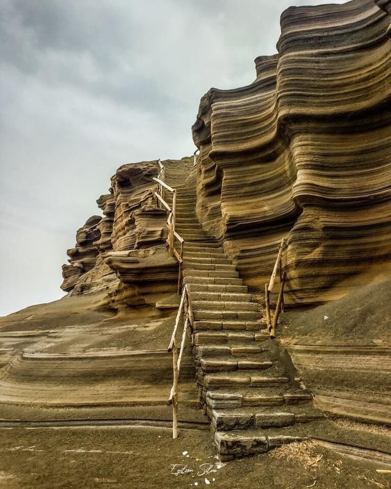 A natural rock formation displays distinct layers, featuring a staircase made of stone steps set against the textured, weathered cliffs. The staircase leads upward, inviting exploration of the scenic landscape above, with a muted sky providing a soft backdrop.
