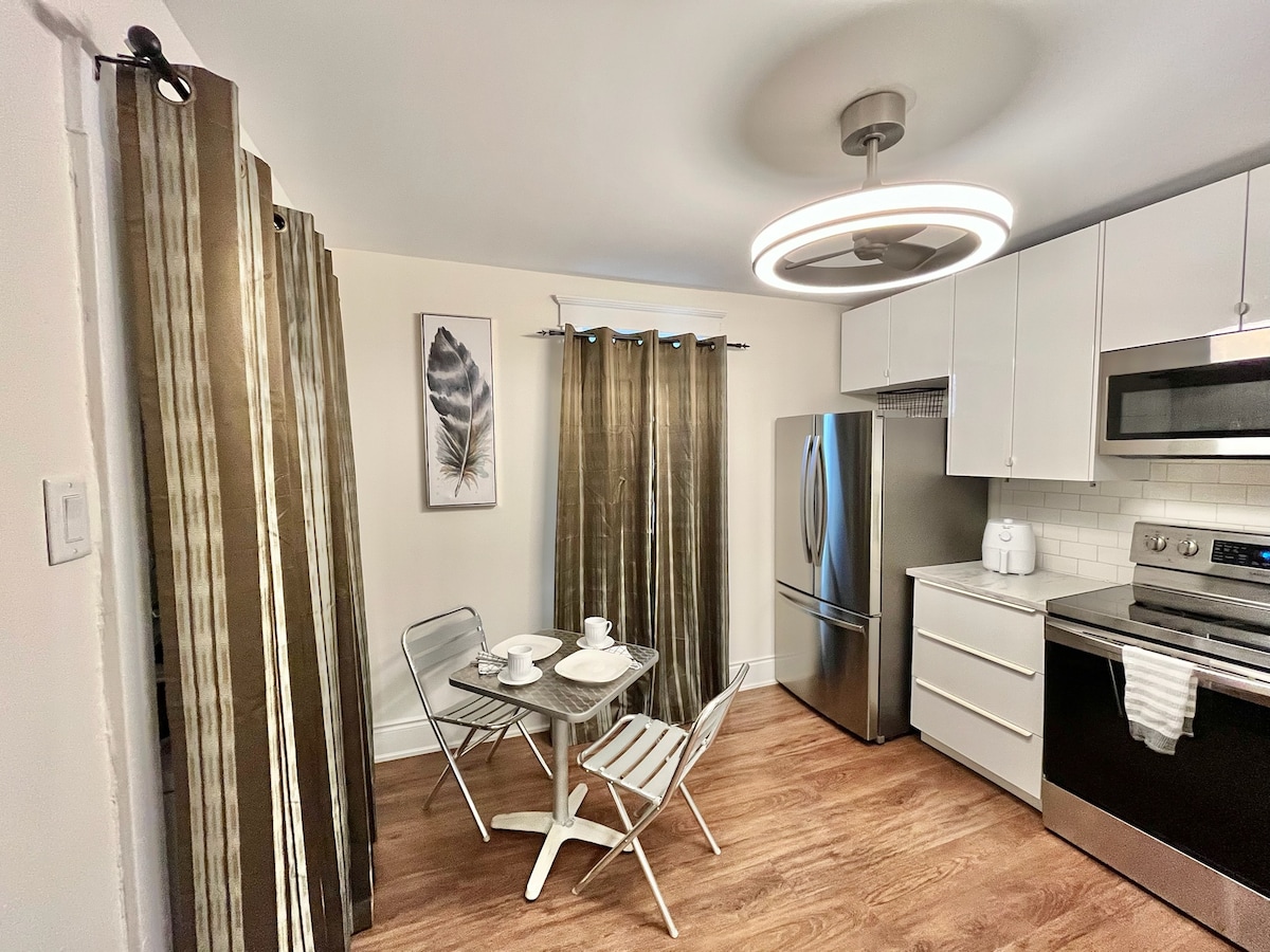 A modern kitchen area features a sleek refrigerator and stainless steel appliances. A round table with two chairs is set with plates and utensils, illuminated by an overhead light fixture. Light fabric curtains frame the window, adding a soft touch to the space.