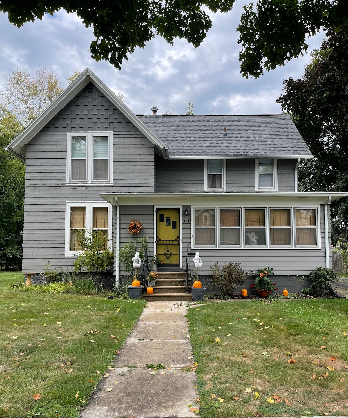 A charming two-story cottage features a gray exterior with textured siding and a gabled roof. A front porch is adorned with seasonal decorations, including pumpkins. Lush green grass surrounds the walkways, leading to the house's entrance. A few trees provide shade in the front yard.
