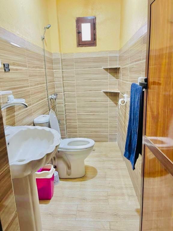 A compact bathroom features beige tile walls and a light-colored floor. A showerhead is mounted above a small tub, and a toilet is positioned next to a light pink trash bin. Two shelves are installed on the wall for convenience, and a small window allows natural light.