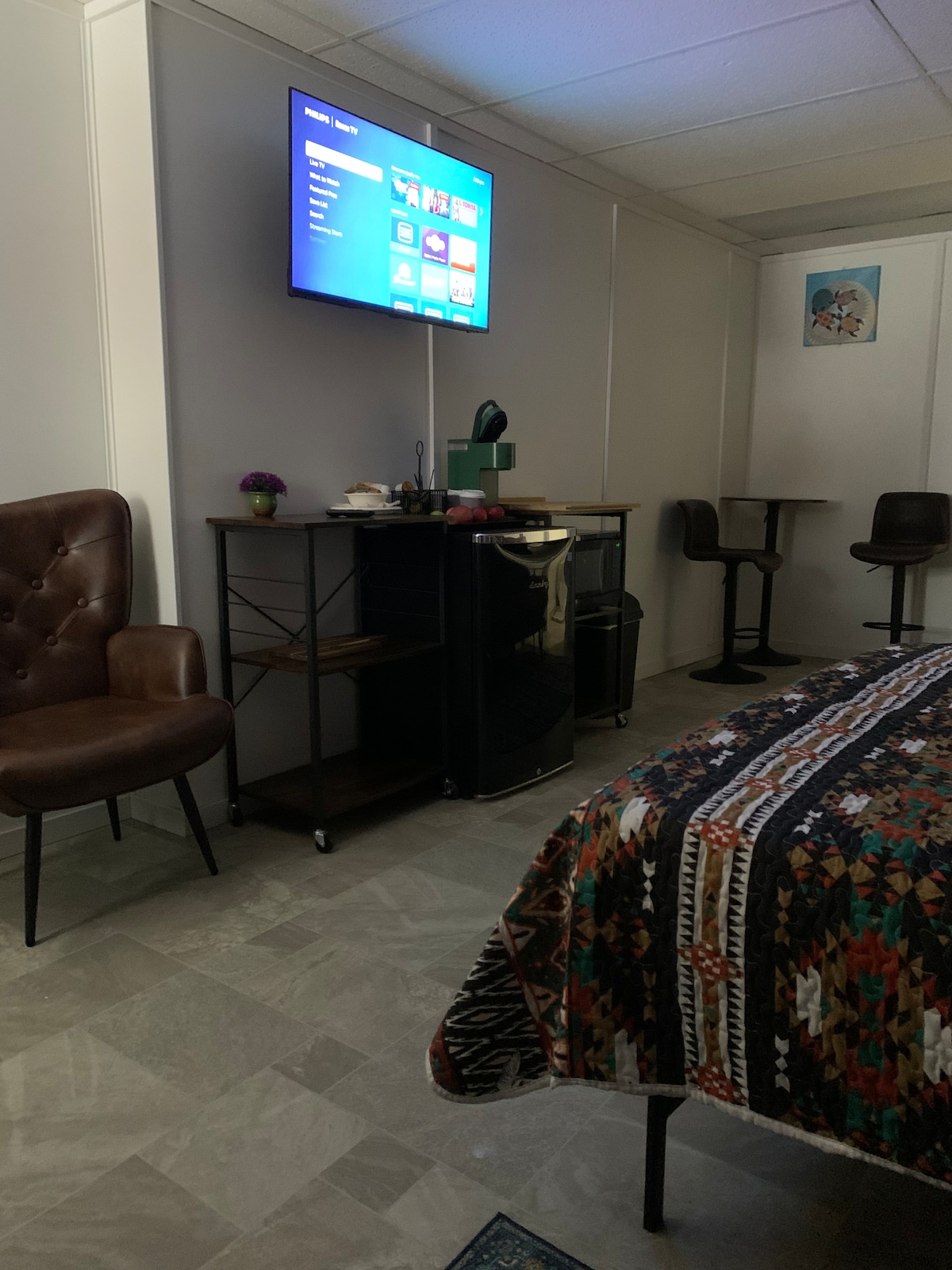 A cozy seating area features a brown armchair and two bar stools at a tall table. A small refrigerator and coffee station are positioned beside a wall-mounted television. A patterned throw blanket covers the spacious bed, with soft, neutral flooring enhancing the overall ambiance.