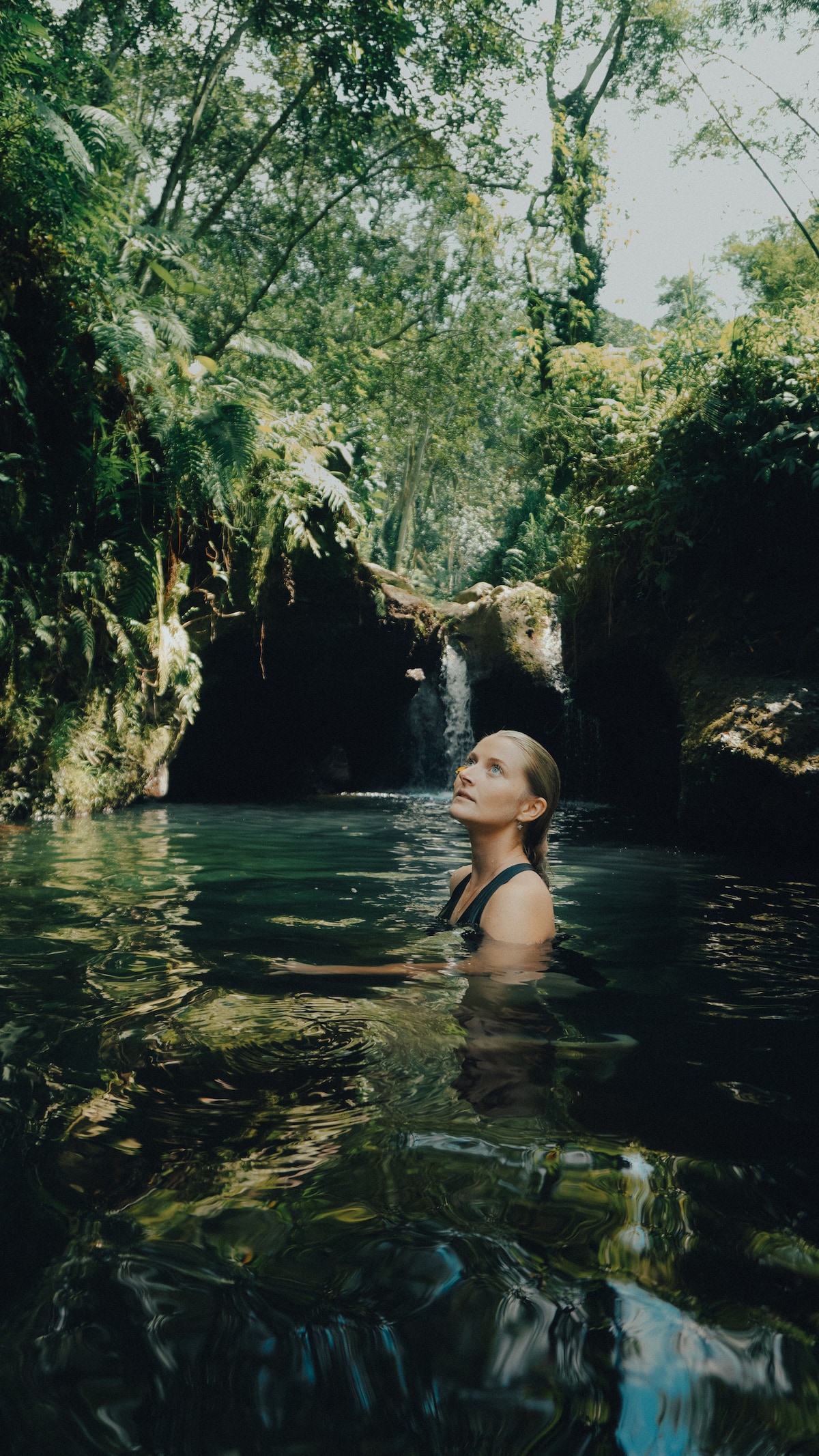 A serene natural pool is surrounded by lush greenery and rock formations. Sunlight filters through the trees, illuminating the calm water. A gentle waterfall is visible in the background, enhancing the tranquil atmosphere of this peaceful retreat.