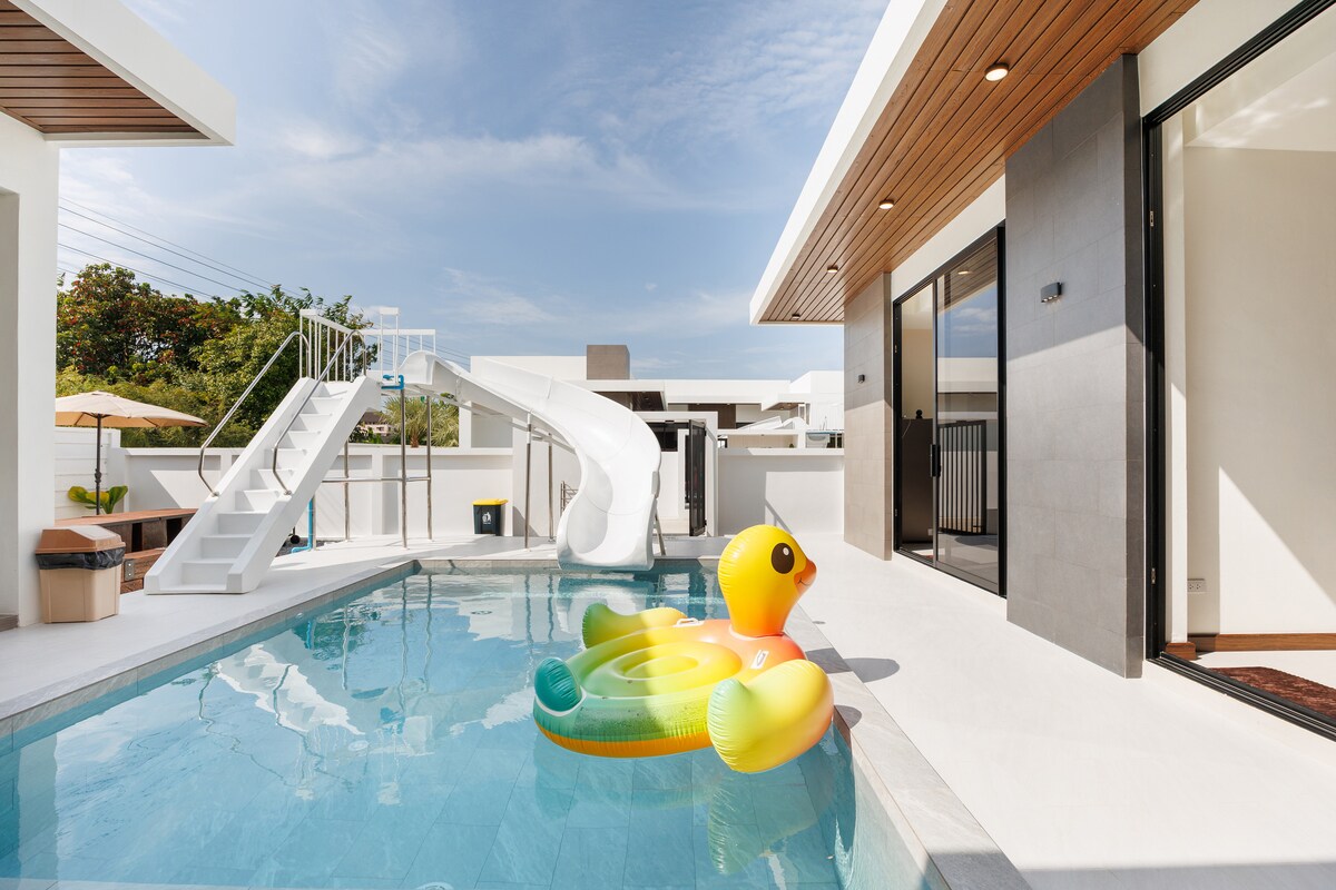 A private pool area features a saltwater pool with a fun waterslide. An inflatable duck float is positioned in the pool, while a sunny patio area is visible nearby, complete with outdoor seating and an umbrella. Modern architecture enhances the inviting outdoor space.