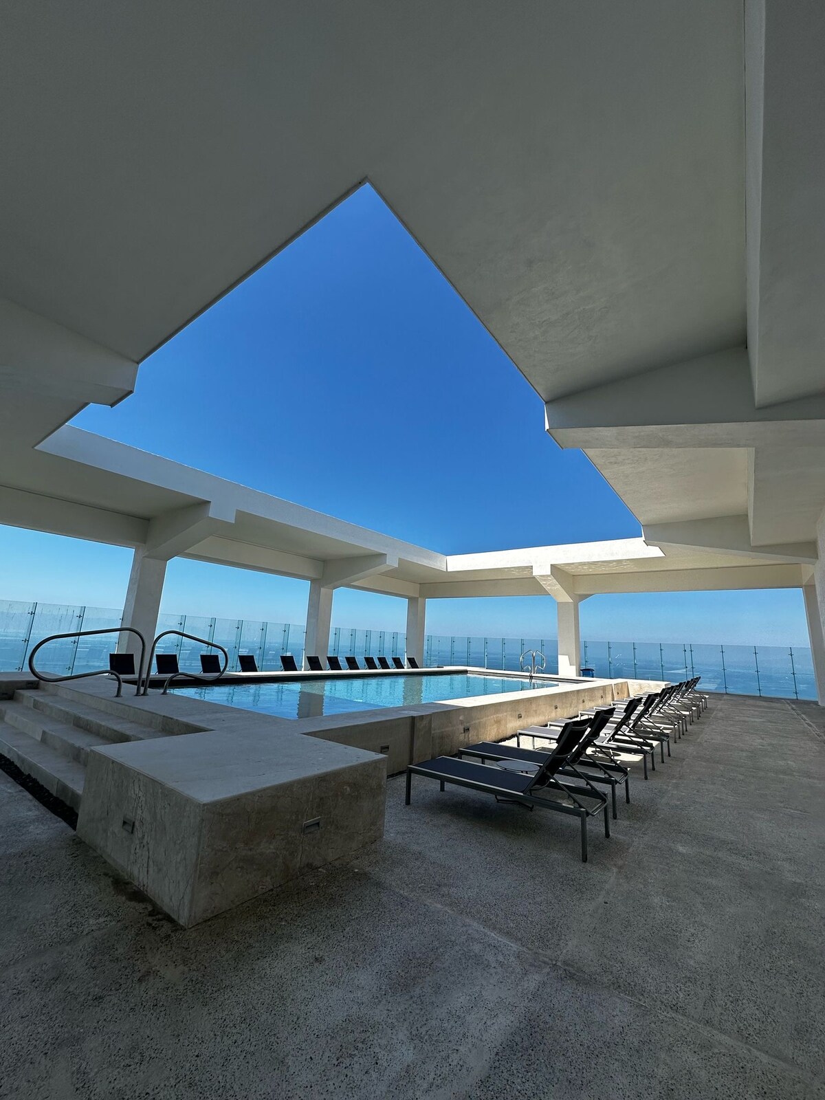 An inviting pool area is situated under a striking architectural overhang, with a clear blue sky visible above. Several lounge chairs line the edge of the pool, providing ample seating. The scene opens up to a vast ocean view, enhancing the atmosphere of relaxation.