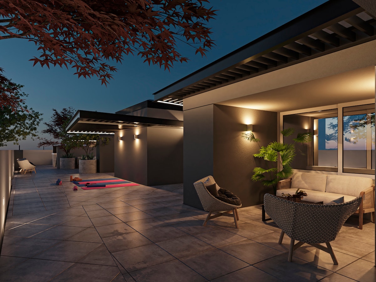 An outdoor terrace features modern seating arrangements, surrounded by greenery and decorative pots. Soft lighting enhances the evening ambiance, while an exercise mat lies on the tiled floor. The view captures an expansive sky, transitioning into dusk.