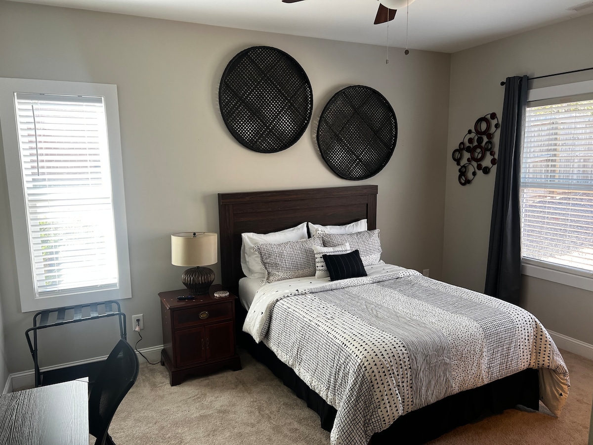A well-furnished room features a cozy bed with fresh linens and decorative pillows, accompanied by a bedside table and lamp. A small desk provides a work area, while two windows allow natural light to brighten the space. Modern wall decor adds character to the room.