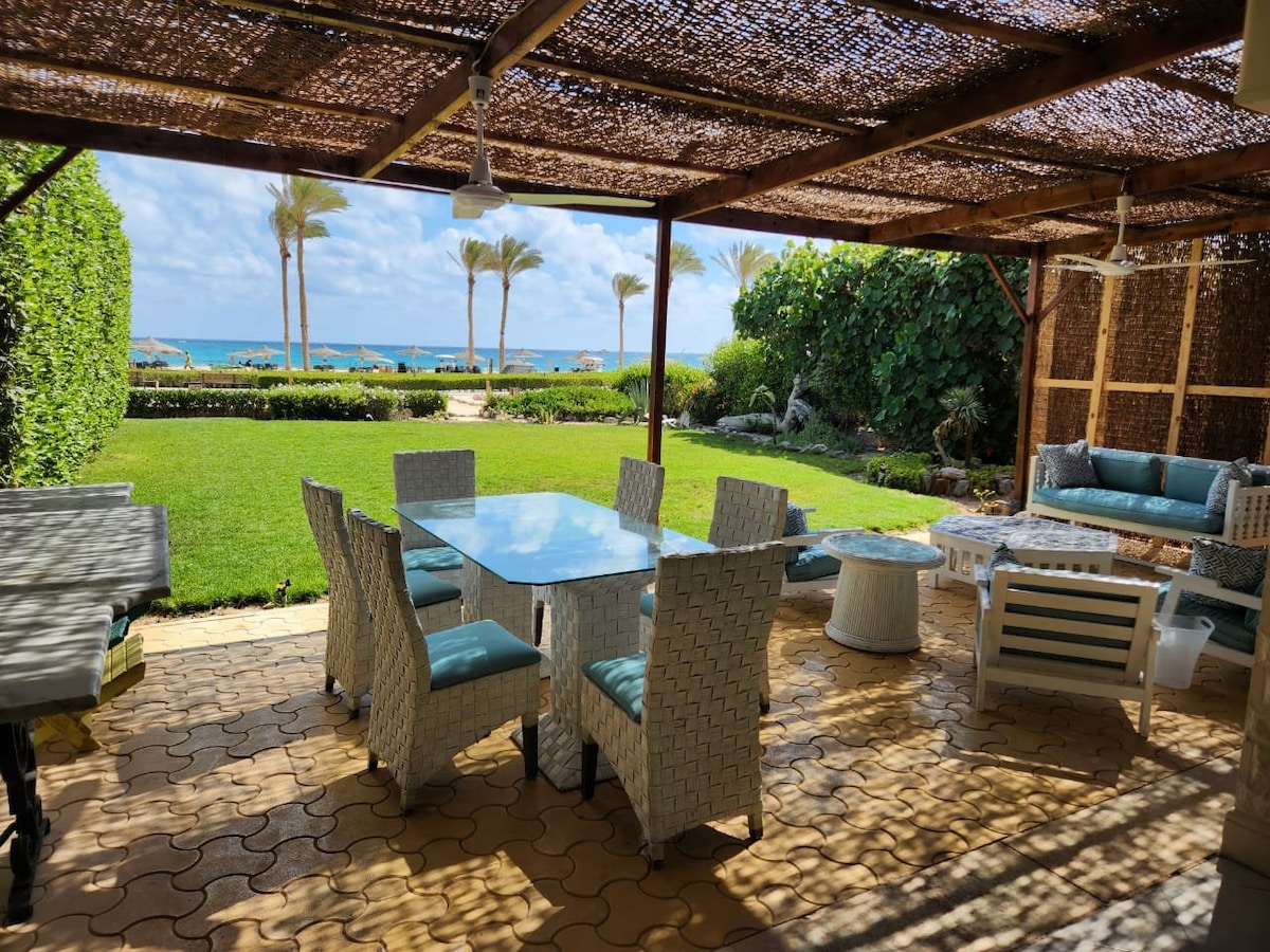 A covered outdoor seating area features a large dining table surrounded by white wicker chairs and soft blue cushions. Lush green grass and palm trees lead towards the beach in the background, with a clear view of the ocean and vibrant sky.