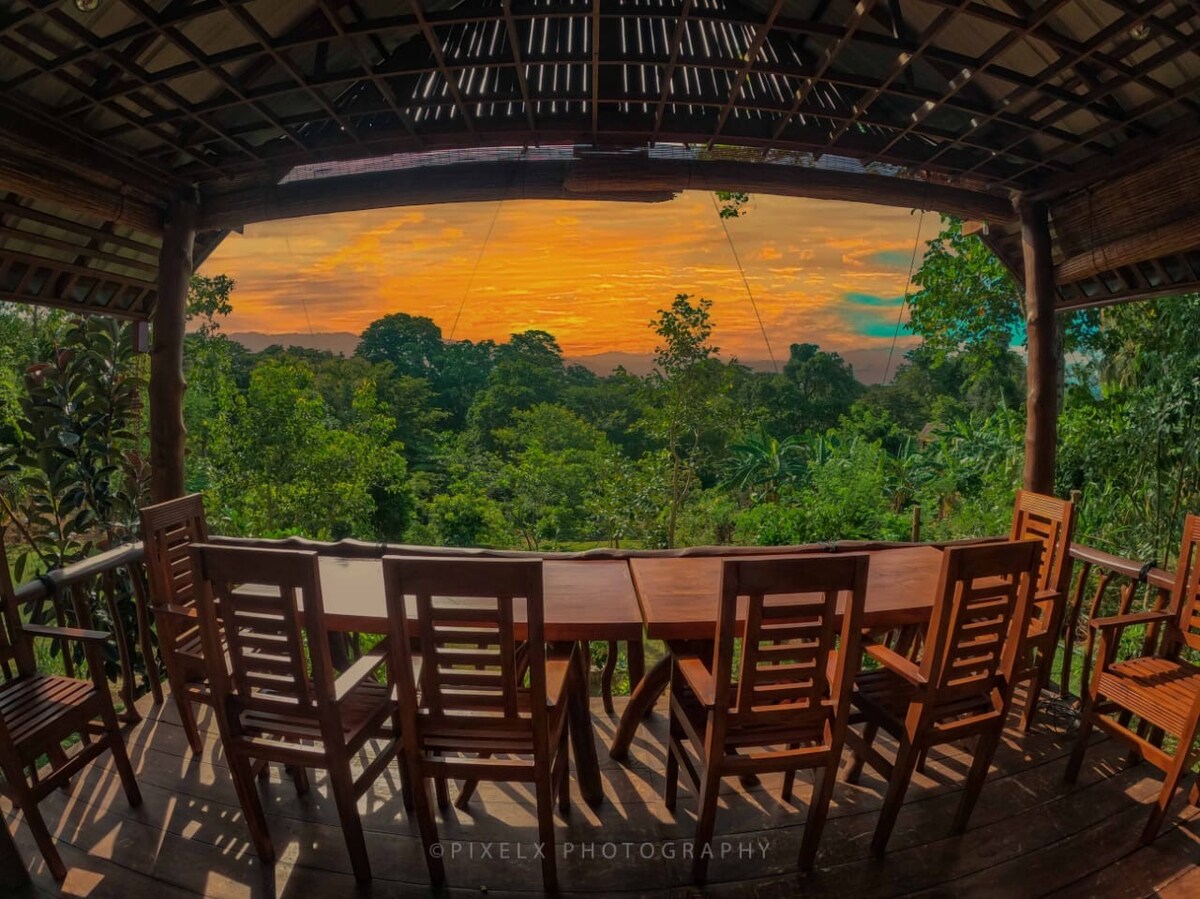 A wooden dining area is positioned on a covered patio, featuring several wooden chairs surrounding a long table. The backdrop showcases a vibrant sunset over lush greenery, creating a serene atmosphere. The space invites relaxation while enjoying nature's beauty.