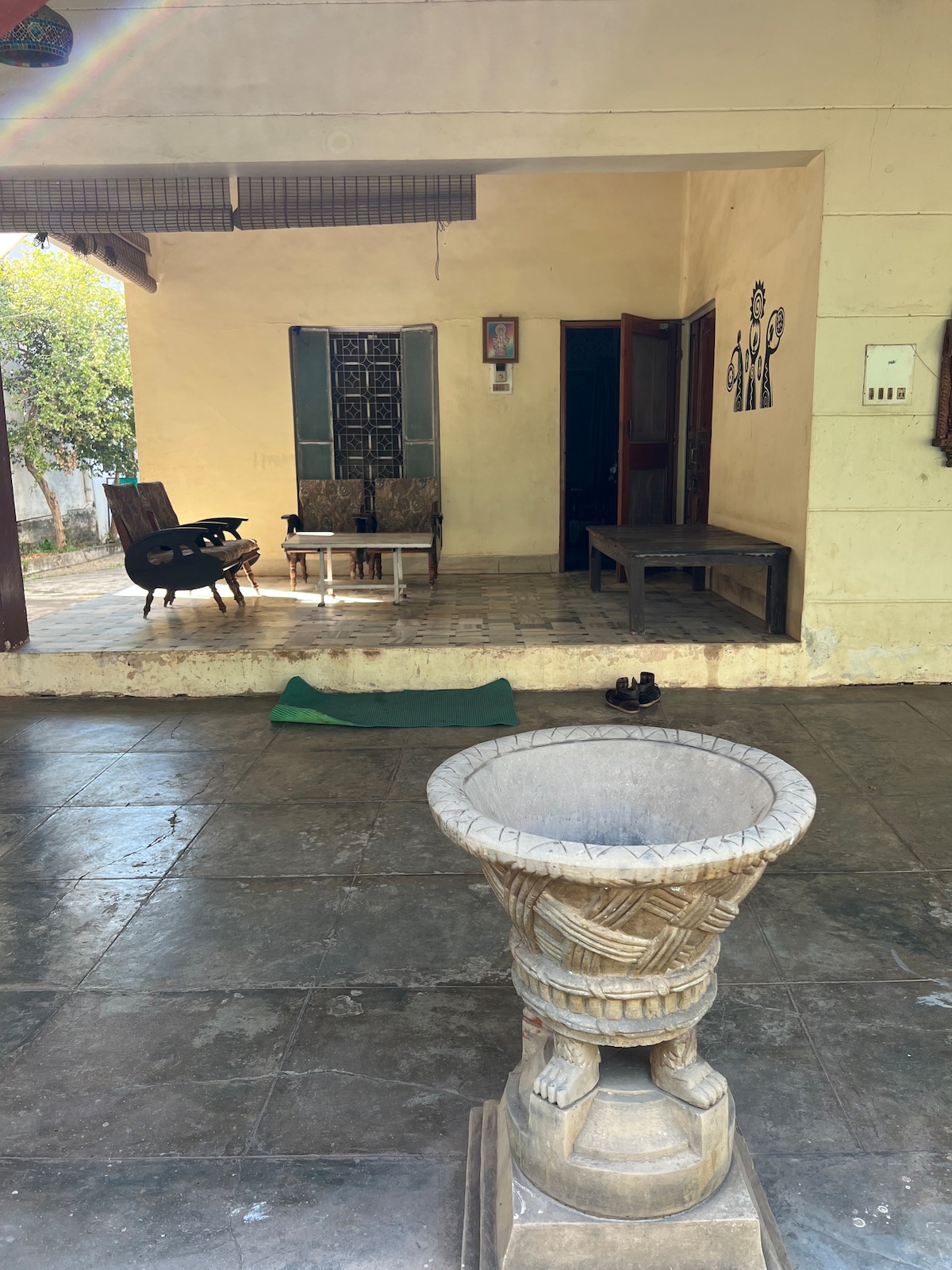 An outdoor patio area is observed, featuring a decorative stone pedestal at the forefront. Two wooden chairs and a table are positioned against the wall, beside a large wooden door leading inside. Light filtering through the patio creates a calm ambiance.