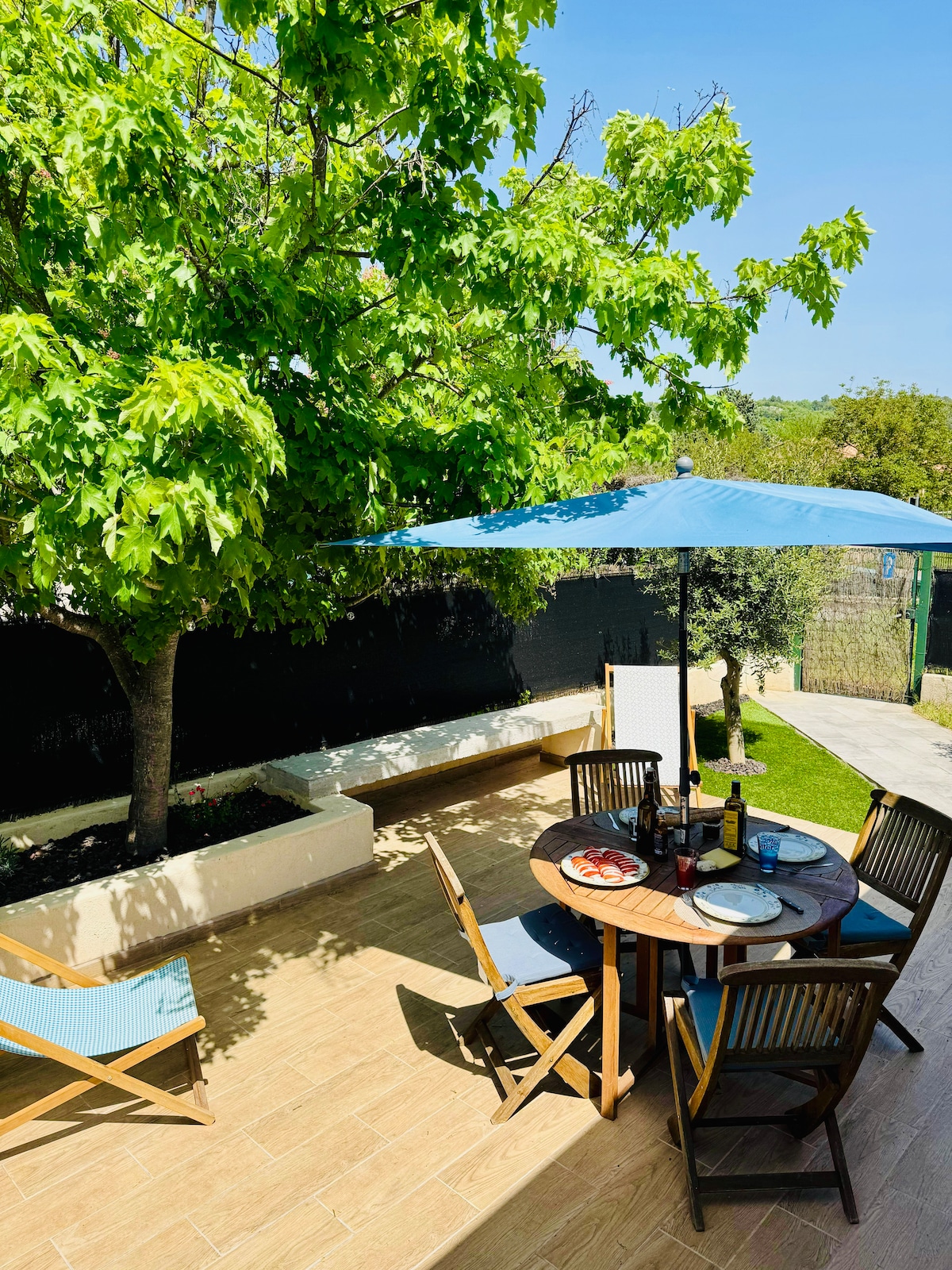 An outdoor dining area is present, featuring a round table surrounded by wooden chairs. A large blue umbrella provides shade, while a tree adds greenery to the space. Lounge chairs are positioned nearby, and a garden is visible in the background.
