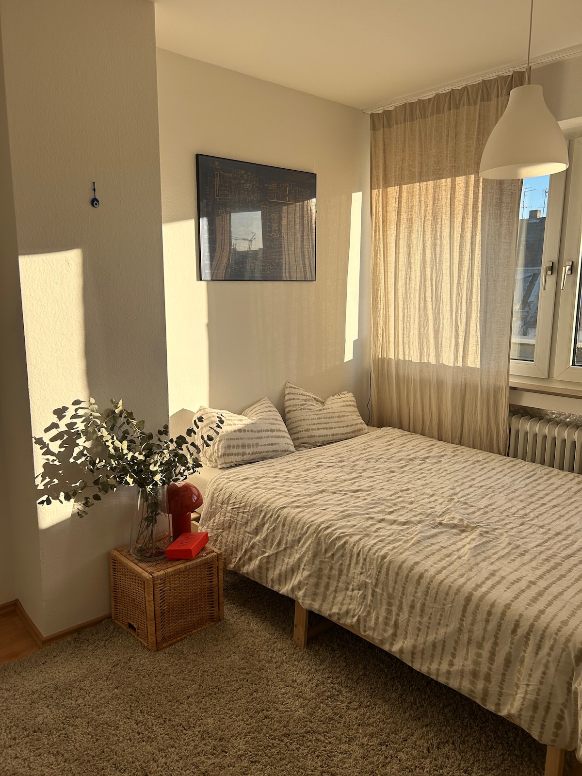 A well-lit bedroom features a spacious bed with a striped blanket and two pillows. A woven basket stands beside the bed, topped with a small red object. Natural light filters through sheer curtains, illuminating the warm tones of the room.
