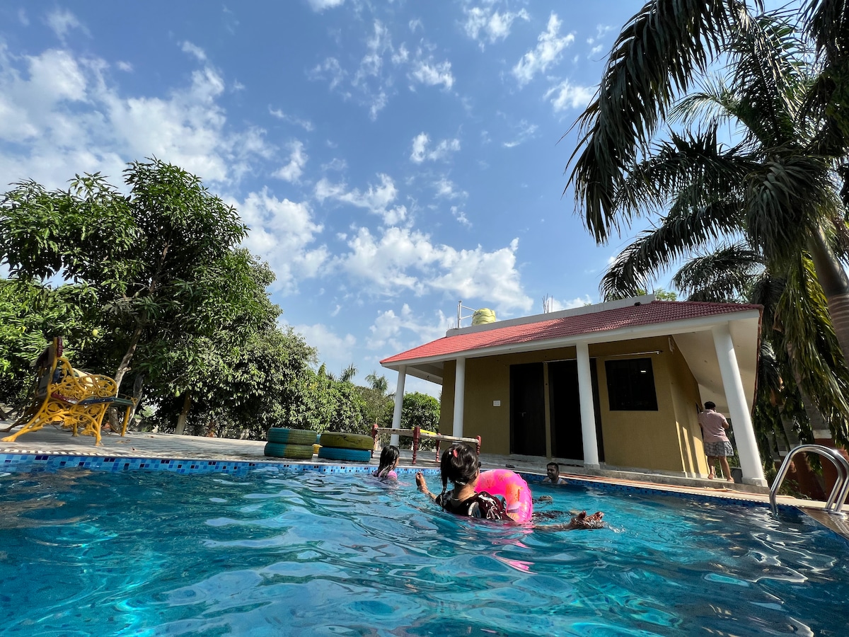 A swimming pool is seen, surrounded by greenery and palm trees. A yellow building with a red roof stands nearby, featuring large windows. Bright blue skies with scattered clouds provide a vibrant backdrop, while individuals enjoy the pool, signifying a relaxing and active atmosphere.
