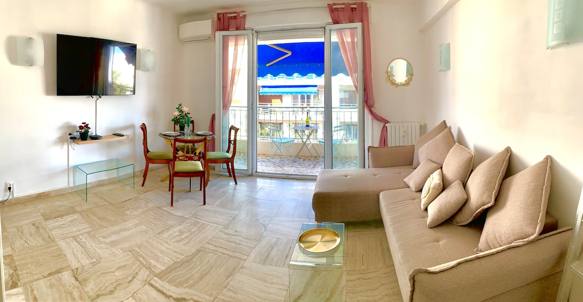 A stylish living area features a large sofa with plush cushions, complemented by a glass dining table and four chairs. Natural light fills the room through sliding glass doors leading to a balcony. Decorative elements include a small round mirror and a potted plant.