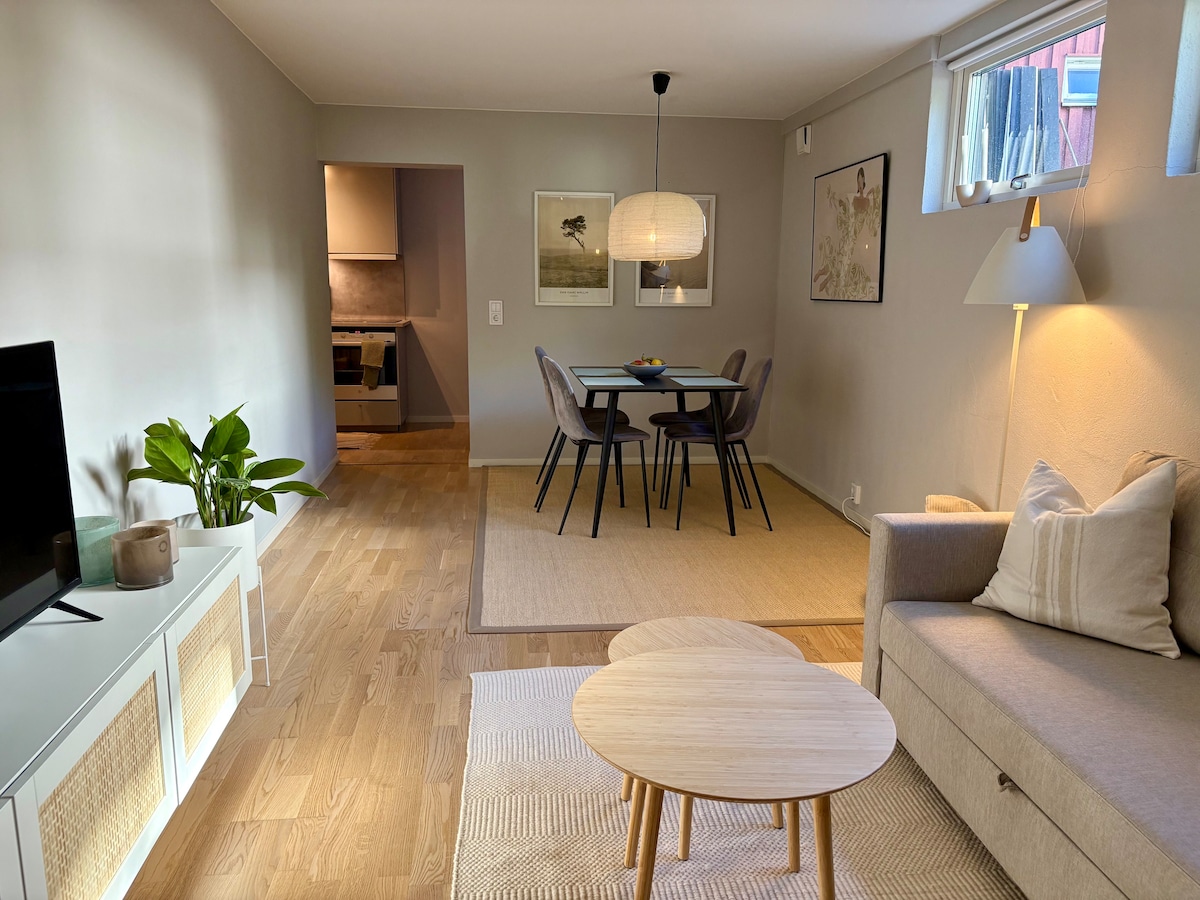 A light and airy living space features a comfortable sofa paired with a round coffee table. A dining area with four chairs is visible, complemented by a pendant light overhead. The open layout connects to a kitchen area, with natural light coming from the windows.
