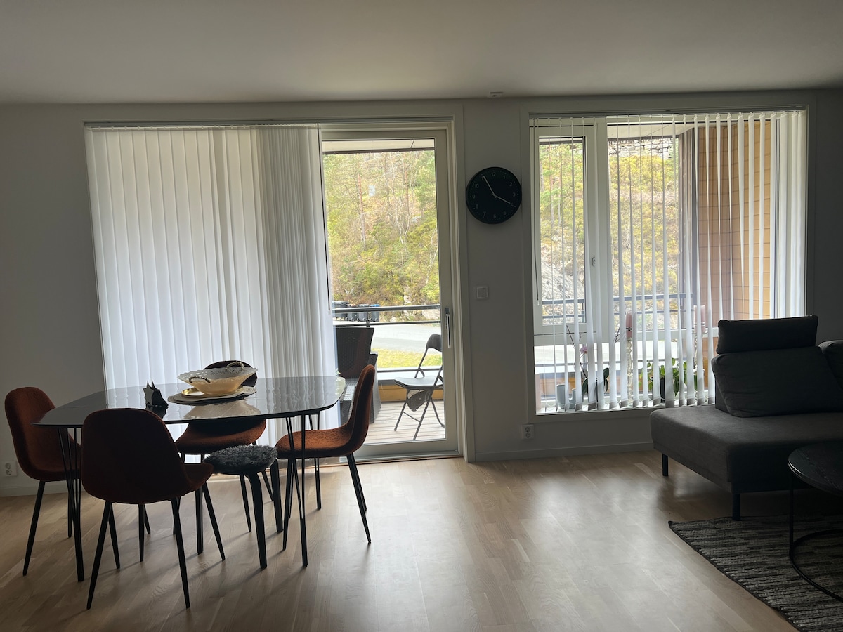 A modern dining area features a round table set with decorative items and surrounded by four chairs. Large sliding glass doors open to a view of the outdoors, while vertical blinds allow natural light to enter the room. A wall clock is visible, indicating the time.