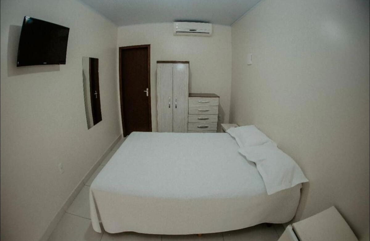 A simple room features a spacious bed dressed in white linens, positioned against a neutral-colored wall. A television is mounted on the wall, and a wooden door leads to another space. A dresser with multiple drawers stands nearby, completing the functional layout.