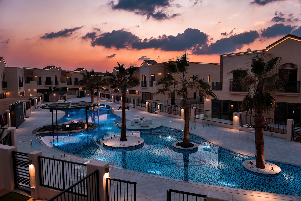 The image captures a serene evening scene of a community swimming pool surrounded by lush palm trees. The pool is illuminated with soft lighting, reflecting the twilight sky. Modern buildings enclose the area, creating a tranquil atmosphere.