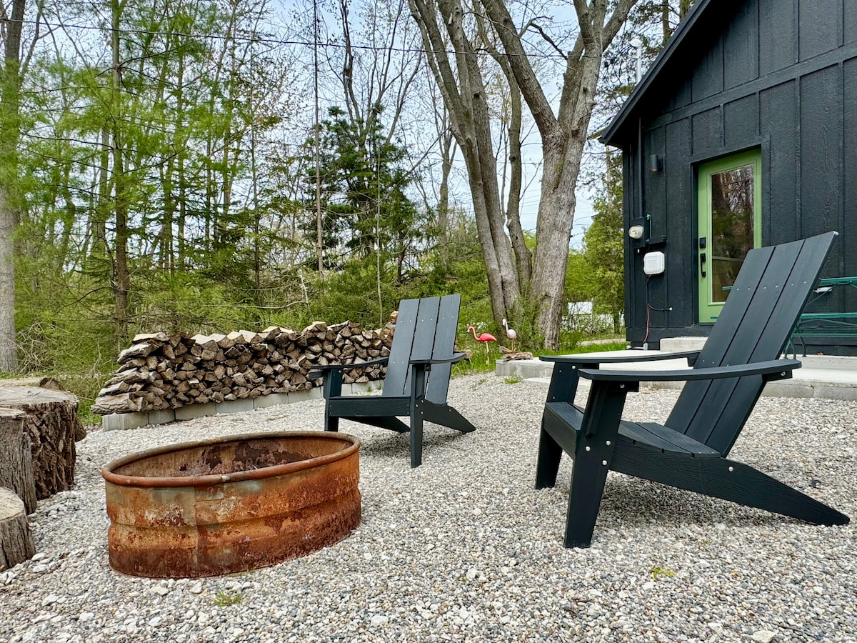 A small patio area features two black Adirondack chairs facing a rustic fire pit, set on a gravel surface. In the background, a stack of firewood leans against a green wall, surrounded by lush trees.